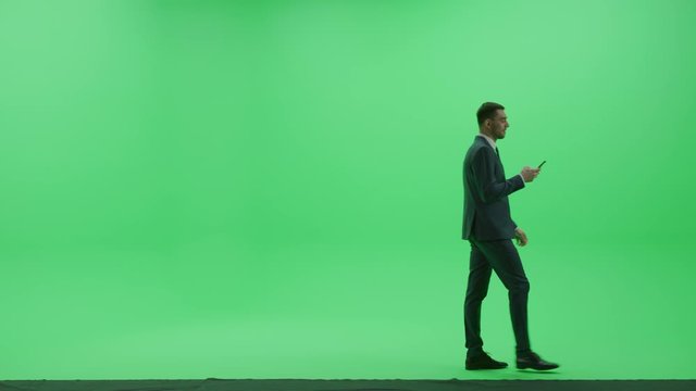 Chroma Key Studio: Handsome Businessman wearing Suit Uses Smartphone to Browse through Internet and Social Media Walks Across the Green Screen Room. Side View Camera Shot