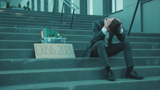 Sad young man sitting on stairs head off with box of personal stuff and poster with the inscription Crisis 2020 got fired due to coronavirus crisis unemployed depression employment economy slow motion