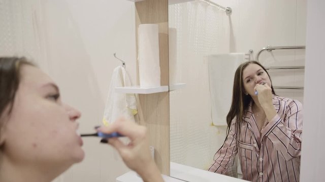 Young girl with long dark hair in a pink nightgown has fun washing and brushing her teeth at home in the bathroom