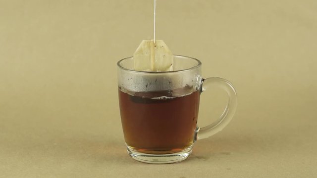 black tea bag lifting and dropping into glass transparent mug to brew tea isolated on beige background slow motion close up .