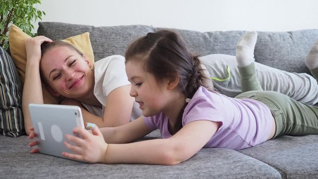 Tired mother yawns and falling asleep while her small daughter using digital tablet on sofa at home. Girl learns technology during Social distance and self isolation in quarantine Coronavirus Covid19