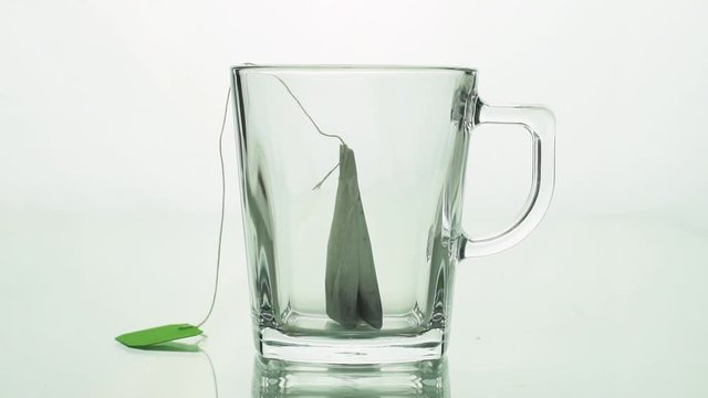 black tea bag with green label is placed in glass transparent mug and filled with boiling water isolated on white background slow motion