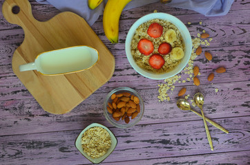 Delicious and healthy homemade milk oatmeal porridge with banana and strawberry and almond nuts, two teaspoons on the lilac kitchen table. Vegetarian or diet food. A hearty snack for energy