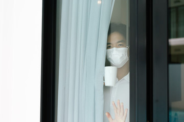 Sick asian male of corona virus looking through the window and wearing mask protection and recovery from the illness in home. Quarantine. Patient isolated to prevent infection.
