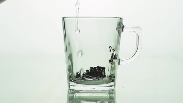 black tea particles pouring into glass transparent mug and filled with boiling water to brew tea isolated on white background slow motion close up
