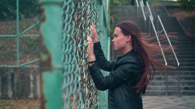 Beautiful and passionate girl in a biker jacket behind an iron fence. Young woman behind bars locked metal fence. Under the jacket, the girl has a burgundy silk top with lace.