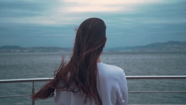 A girl with long hair in a white sweatshirt stands with his back in the frame against the backdrop of a stormy sea. A strong wind is blowing. Cloudy day, the sea in a storm, big waves.