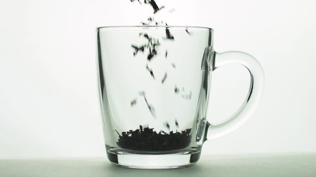 black tea particles pouring into glass transparent mug and adding boiling water to brew tea isolated on white background slow motion close up
