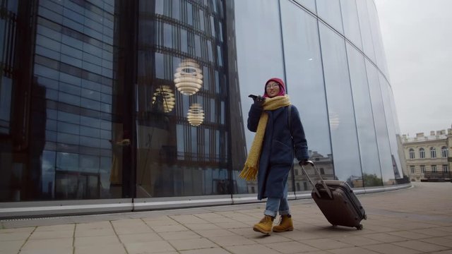 Young asian female tourist walking with luggage in city downtown, smiling and chatting via speaker phone while looking for hotel after arrival