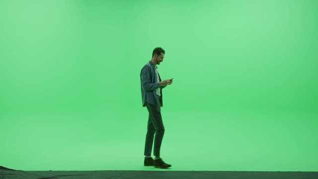 Green Screen Studio: Strong Handsome Man Wearing Casual Clothes, Uses Smartphone Device, Talking on the Mobile Phone, talking to a Girlfriend Walks Across Chroma Key Room. Side View Camera Shot