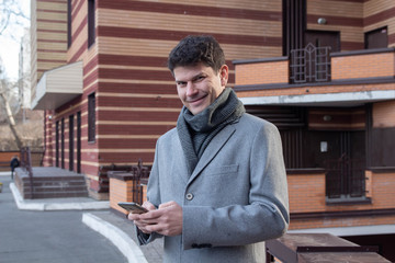 Obraz premium positive man in long coat holds digital gadget posing and smiling against urban building on street. Concept fashion city travelling