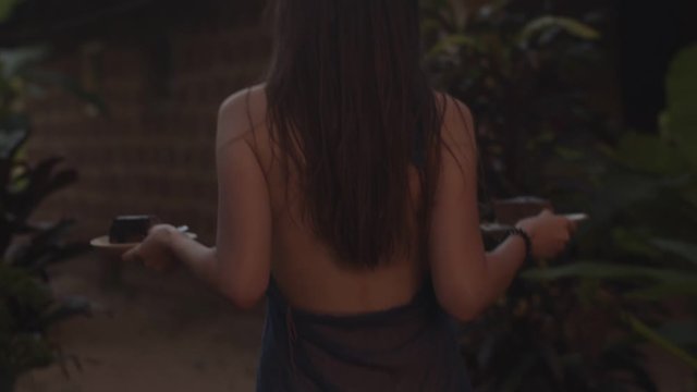 Back view of unrecognizable woman carrying plates with food outdoors natural twilight light. Faceless elegant girl in dress with no back going outside slow motion. Female beauty gracefulness concept