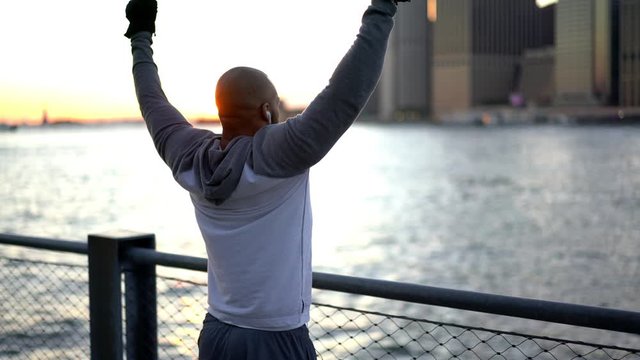 Back view of motivated dark skinned athlete in tracksuit raising hands in air celebrating sport triumph and goal, accomplished bodybuilder listening music via electronic headphones
