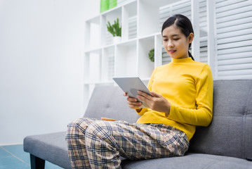 Young asian girl happy woman sitting on sofa watching movie with tablet at home. After being detained at home due to the outbreak of coronavirus covid-19.