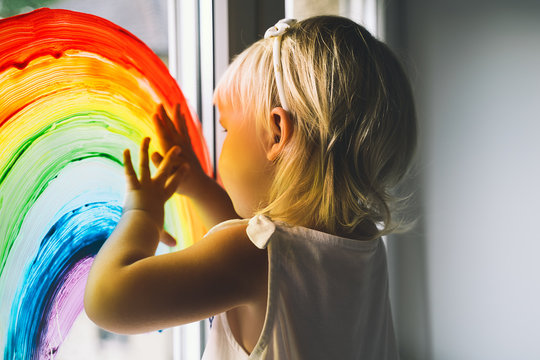 Little girl hands touch painting rainbow on window.
