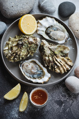 Fresh opened iced oysters on a round serving plate, vertical shot on a dark-grey stone background with pebbles