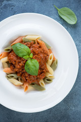 White plate with three-colored penne and bolognese sauce, vertical shot on a blue stone background, above view