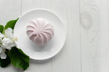 Pink marshmallows on a white porcelain plate Branch with white flowers of apple trees on white wooden background