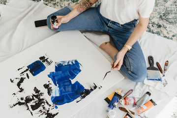 Woman's hand drawing on white canvas using phantom blue and black oil colors. Many drawing instruments near on the floor.