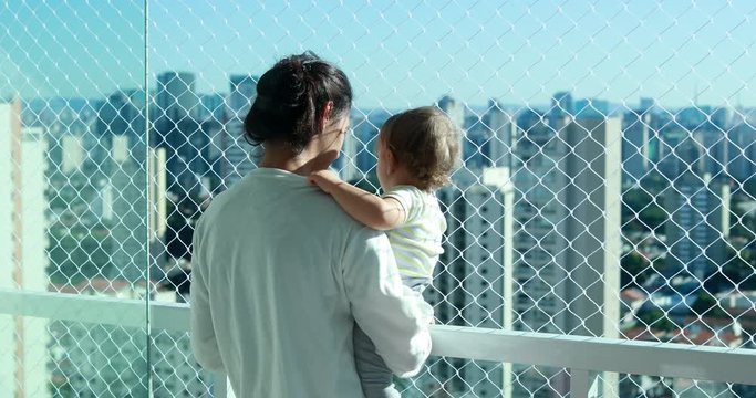 Mother holding baby in arm looking at metropolitan city view. Mom and infant in apartment balcony with safety net