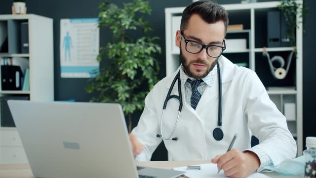 Young man doctor in white gown working with laptop computer and writing sitting at desk in office. Modern technology, medics and medicine concept.