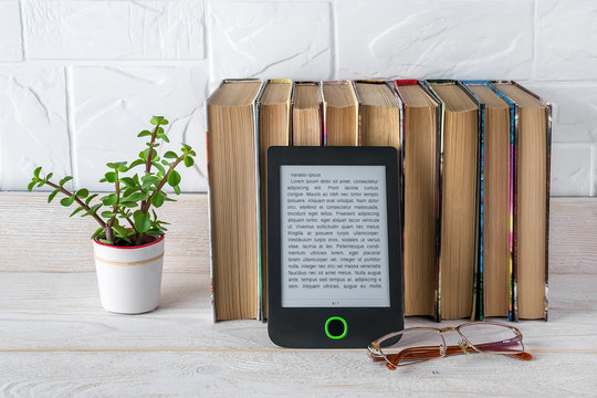 E-reader near stack of paper books, potted plant and vision glasses on a white wooden table. Copy space on e-book display. E-reading for pleasure and education.