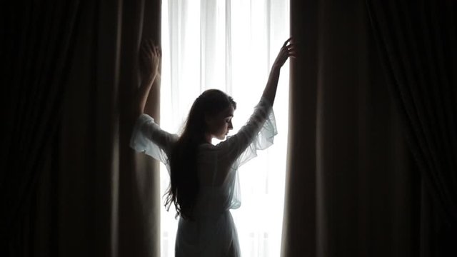 Young woman raises her hands and runs them over curtains in bedroom, shielding herself from morning light that comes in through the window. Silhouette of beautiful girl opens the curtain on the window