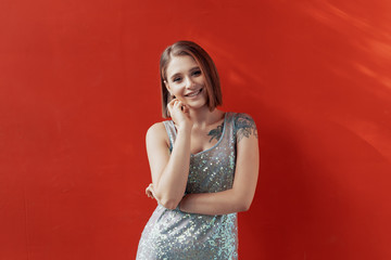 Cheerful young girl posing in front of a red wall. Natural light. Tattoo on left shoulder. Dressed in a shiny dress