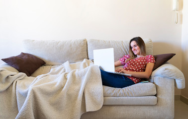 Woman sitting on the sofa while watching a laptop and online learning and teleworking from home while smiling.