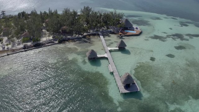 Aerial shot of huts on pier over sea by island during sunny day, drone flying over water - Coco Plum Cay, Belize