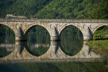 Fototapeta na wymiar The Ottoman Mehmed Pasa Sokolovic Bridge in Visegrad, Bosnian mountains, with fantastic river reflection, fisherman on a boat. Bosnia and Herzegovina.