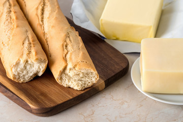 Close-up of fresh wheat baguette on cutting board, cheese on saucer and butter on the kitchen table.  Ingredients for sandwiches, toasts, healthy breakfast or snack.