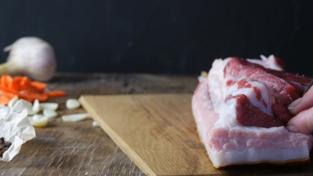 Female hands stabs with a knife and stuffed seasonings with slices of garlic and carrots a piece of fresh raw meat pork steak or bacon in a rustic style on wooden kitchen board