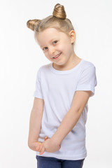 A little blonde girl with a funny hairdo and brown eyes shy smiles and gestures with her hands. White t-shirt, white isolated background, soft light.