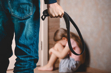 A man with a belt in his hand threatens a little girl. A little girl sits in a corner and hides her face . The concept of domestic violence against a child.