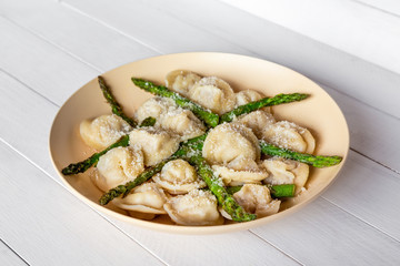 Meat dumplings with parmigiano cheese and fried asparagus on biege plate, on white wooden table, side view, selective focus