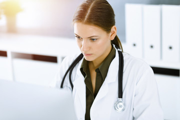 Woman-doctor typing on pc computer while sitting at the desk in sunny hospital office. Data in medicine and healthcare