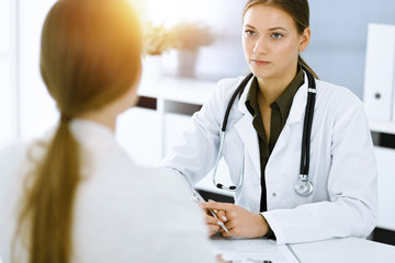 Woman-doctor and patient sitting and talking at sunny hospital office. Green color blouse suits to...
