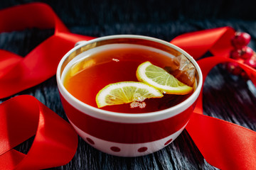 A mug of black tea, a beautiful brown and maroon color, from a disposable bag with a label on a black textured background with a red satin ribbon, lemon slices and berries