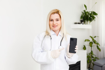 Side profile portrait smiling female doctor, healthcare professional in white lab coat with stethoscope, analyzing data results on mobile smart phone standing in hospital hallway corridor