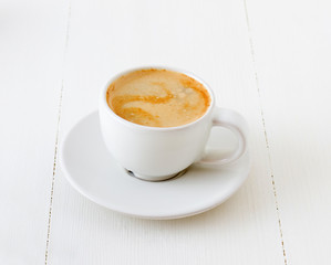 Cup of fresh coffee or espresso with foam in the white porcelain cup and plate. White wooden table background.
