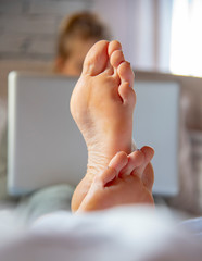 Focus on the feet in the foreground. A teenage girl is lying on a bed with a laptop.