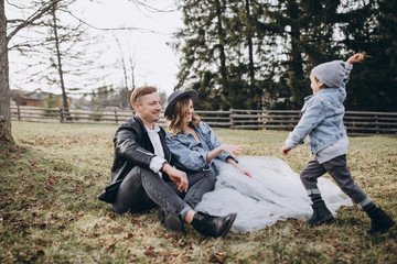 Stylish family in the autumn forest. A guy in a leather jacket and a young girl in a gray-blue dress play with his son in a meadow of a pine forest in the mountains against the background of a fence 