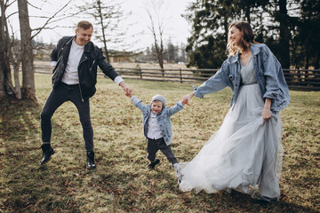 Stylish family in the autumn forest. A guy in a leather jacket and a young girl in a gray-blue dress play with his son in a meadow of a pine forest in the mountains against the background of a fence 