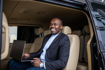 African successful businessman working at a laptop with a grin sitting in the beige interior of a...