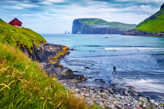 Tourist on a dlack sand beach in Tjornuvik village. Picturesque morning scene of Streymoy island with Eidiskollur cliffs on background. Impressive seascape of Atlantic ocean, Faroe Islands, Denmark.