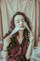 A beautiful young woman with curvy hair holding brushes on the face. A girl in a burgundy linen dress stained with clay. A ceramist in a process of making ceramics in her workshop