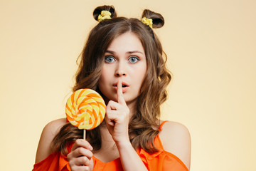 portrait of cute beautiful girl with a round caramel shows gesture shh on a colored background, pretty girl with candy, concept of dieting, secret temptation