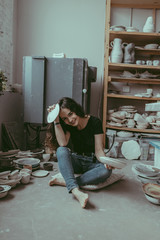 A smiling european young woman sitting on the concrete floor and holding in hands craft made cly dishes of white color. A ceramist at work concept. A various ceramic tableware on a background.