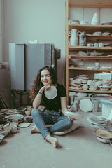 A smiling barefoot young woman sitting on a floor with craft ceramic plates in her hands and surrounded by various tableware as dishes, cups, mugs, vases, bowls, jugs. A ceramist at work concept